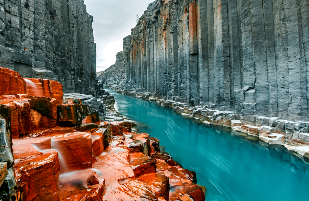 Looking down a rocky canyon in Iceland. The rock columns are a gray color with orange iron stones throughout it. Running through the canyon is a crystal blue river. One of the best stops on an Iceland road trip