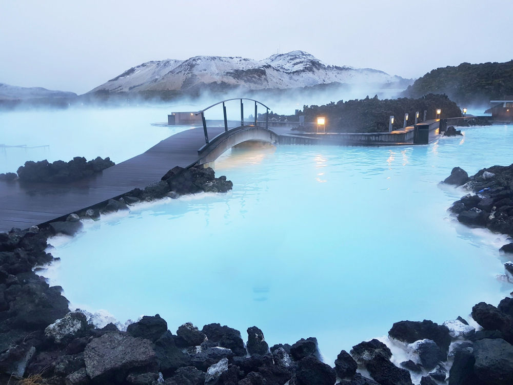 A bridge and walk way with a black railing. It goes over a turquois blue lagoon and there are some black rocks near it. There are lights that light up a path and in the distance is a mountain peak covered in snow. 