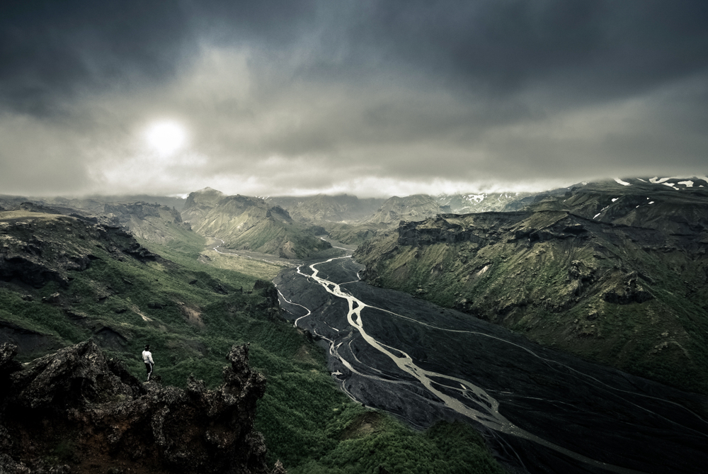 An aerial view of a rocky canyon with a volcanic river bed running through it. It is a dark and moody image with the canyon covered in dark moss and grass and the sky is cloudy. 