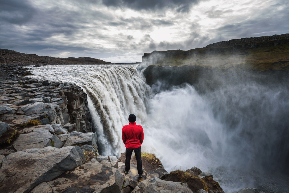 a man in a red jacket, black pants and black hat standing at the edge of a large waterfall with mist rising on a cloudy evening during spring in iceland