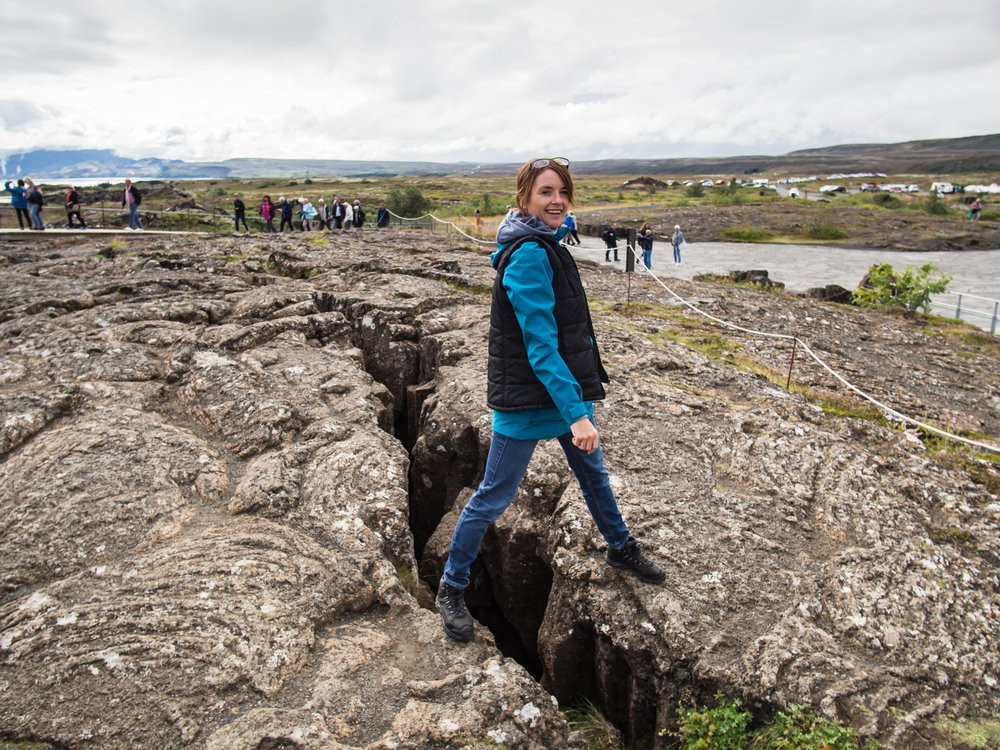 A woman standing across an opening in the a rock formation that is actually where two continental tectonic plates meet. She is wearing jeans, a blue shirt, and a black vest. One of the coolest things to do in Thingvellir National Park. 