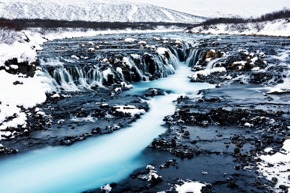 The blue water of Bruarfoss Waterfall flowing between black rocks and snow.