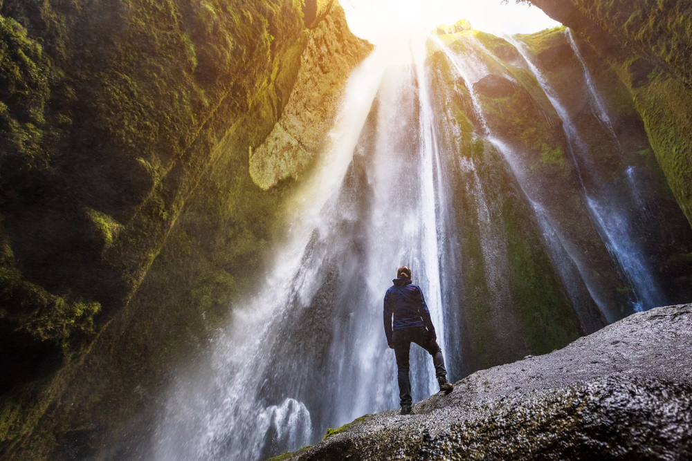 Gljufrabui Canyon in iceland with a hiker standing in front of waterfall falling from above and sunlight shining through 