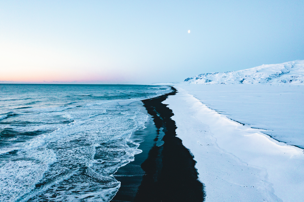 An aerial view of Reynisfjara Beach with the black sand cutting a contrasted line in between the ocean and snow.