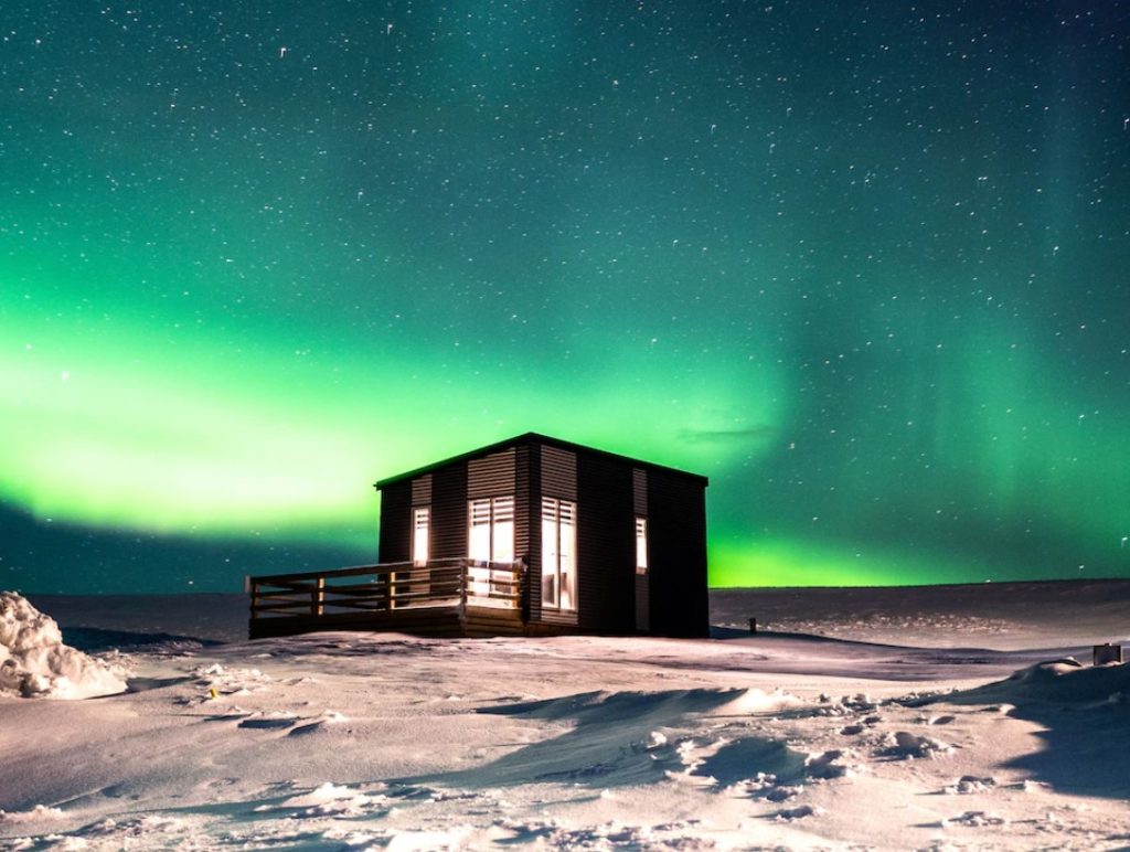 A black cottage lit up from the inside with long narrow windows on the front. It is sitting in a field full of snow and behind it you can see the Northern Lights. One of the best vacation rentals in Iceland. 