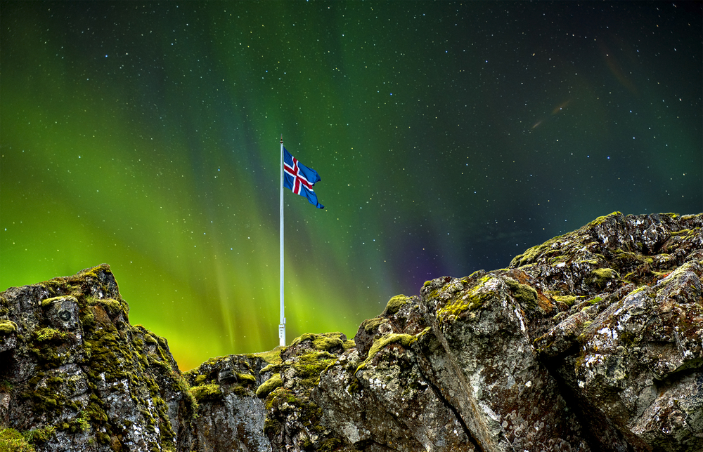 The Icelandic flag sticking out of a large rock formation. It is night time and in the sky you can see the aurora borealis. 