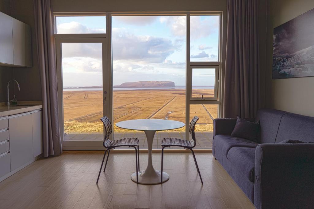 The view from floor to ceiling windows in a cottage in Vik Iceland. In the room you can see part of a kitchen counter, a couch, and directly in front of the windows is a small table with two chairs. Out the windows you can see a straw-colored field that goes on for miles before hitting the ocean. There is a large hill or mountain in the ocean water. One of the best vacation rentals in Iceland. 