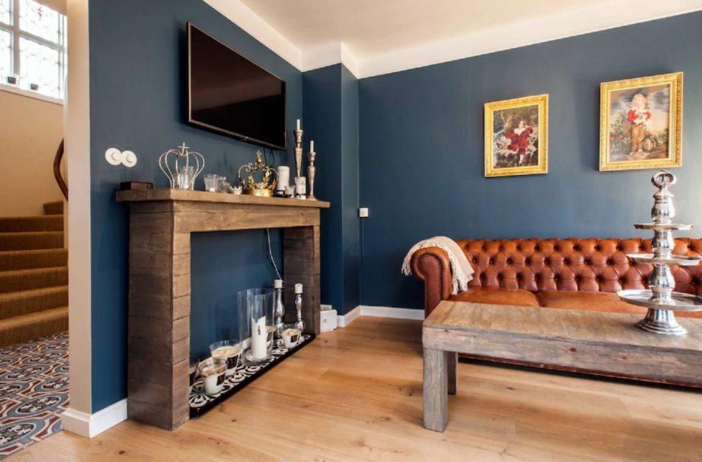 The cozy living room of one of the best vacation rentals in Iceland in central Reykjavik. The room has dark blue walls,  with paintings in ornate gold frames, a brown leather sofa, a mantel filled with candles, a wood coffee table and a tv. 