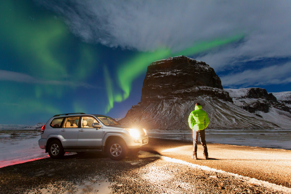 car lights lighting up a person who is watching the Northern Lights dance over some snow covered mountains