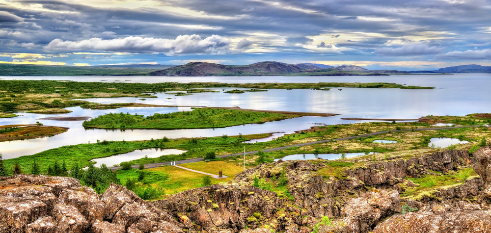 From a vantage point on a rock formation looking out at a large lake in Iceland. You can see small islands in the lake, mountains in the distance, and trails. One of the best things to do in Thingvellir National Park.