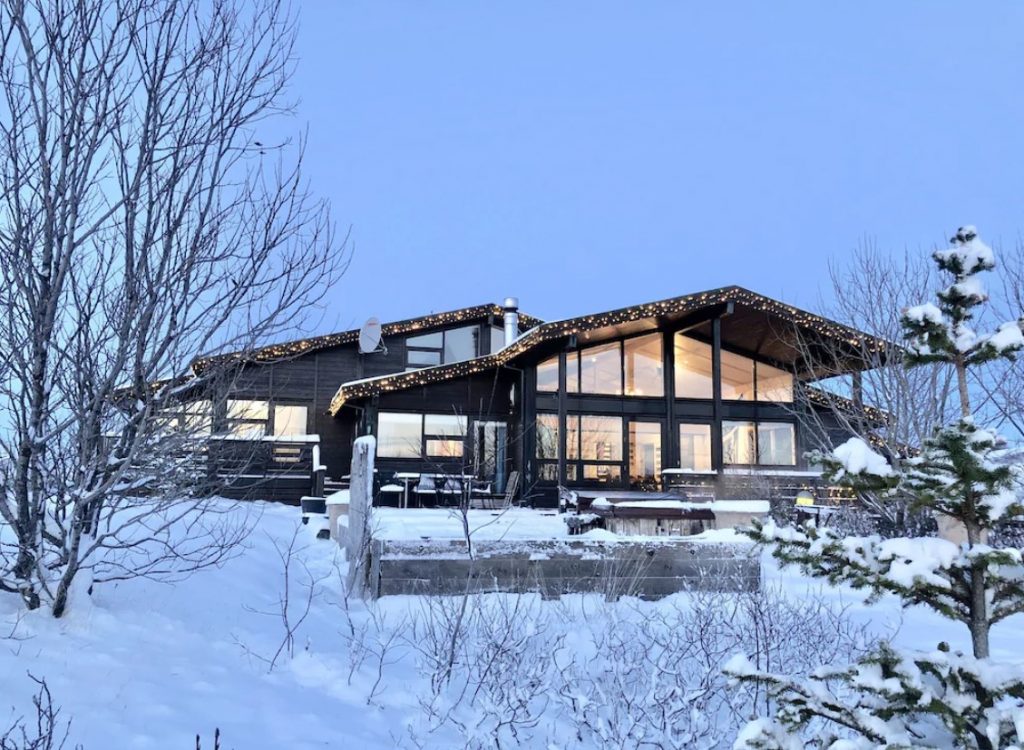 The exterior of a black luxury villa in Iceland. It has large windows, twinkle lights on the edge of the roof, and a large patio out front. The entire villa and landscape is covered in deep snow. 