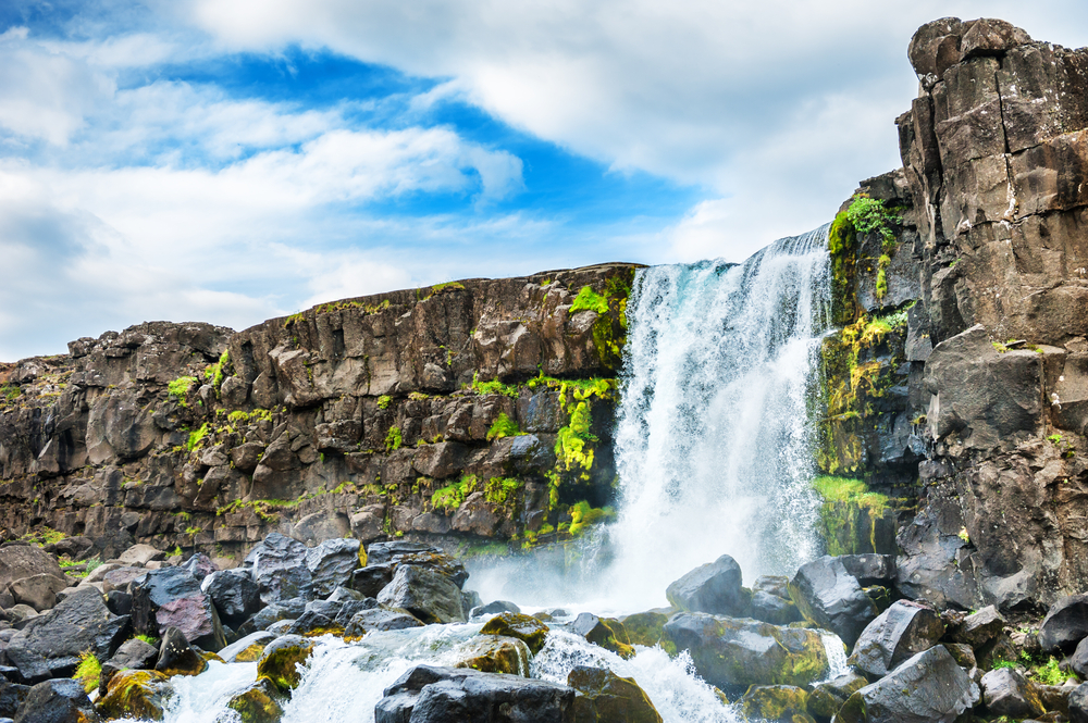 A cascading waterfall over a large rock formation into a shallow river and pool. There is moss and ferns growing on the rocks. One of the best things to do in Thingvellir National Park. 