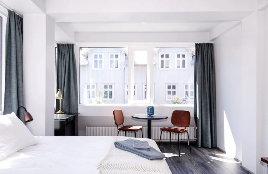 A modern bedroom in a studio apartment. The walls are white and there are large windows that have grey curtains on the sides of them. You can see white buildings outside of the windows. The room has a white bed with a copper lamp next to it, a small table with two wooden chairs, and a desk with a gold lamp. One of the best vacation rentals in Iceland.