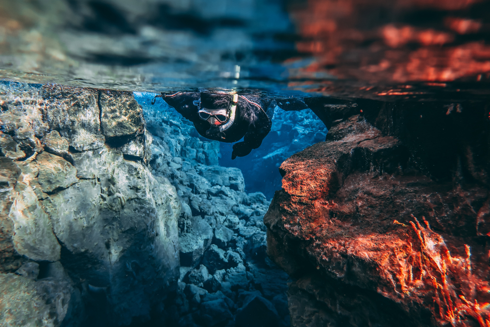 A person in scuba gear snorkeling in the Silfra Fissure. The water is crystal clear and you can see rock formations that appear gray, red, and blue. 