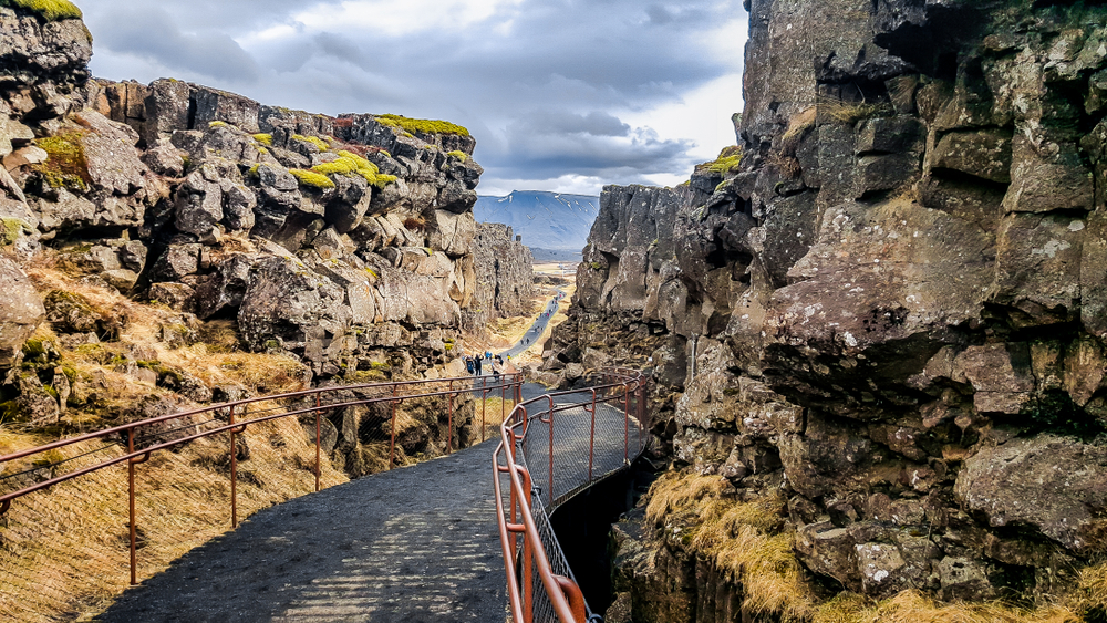 Looking down a black path between two tectonic plates that are sticking up from the Earth's crust. The trail has red guard rails and in the distance you can see people on it. One of the best things to do in Thingvellir National Park. 