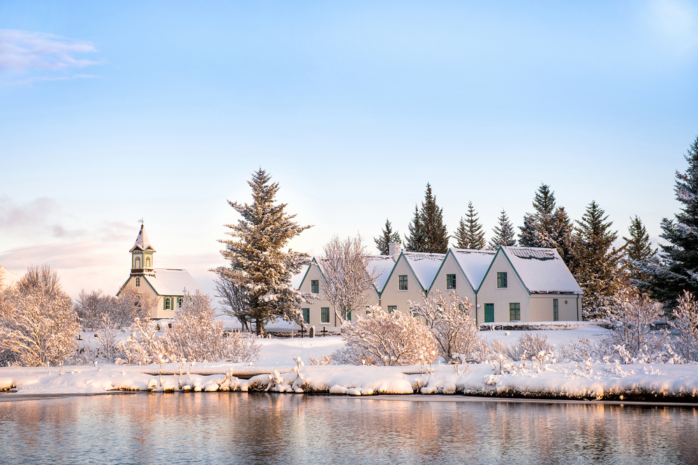 Looking across a lake in the winter at a row of homes connected and an old church. The area is covered in snow in Thingvellir National Park. 