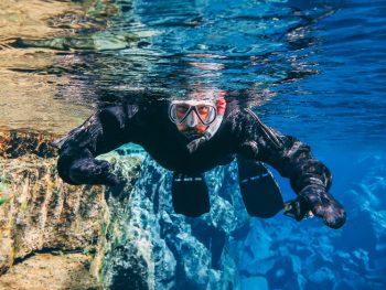 Man in a snorkel mask swims toward camera in the blue waters of the Silfra fissure