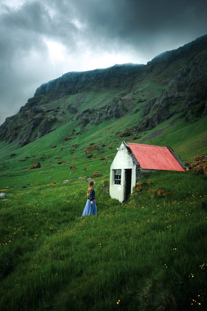 Victoria approaches what appears to be an abandoned building or shed in a grassy field