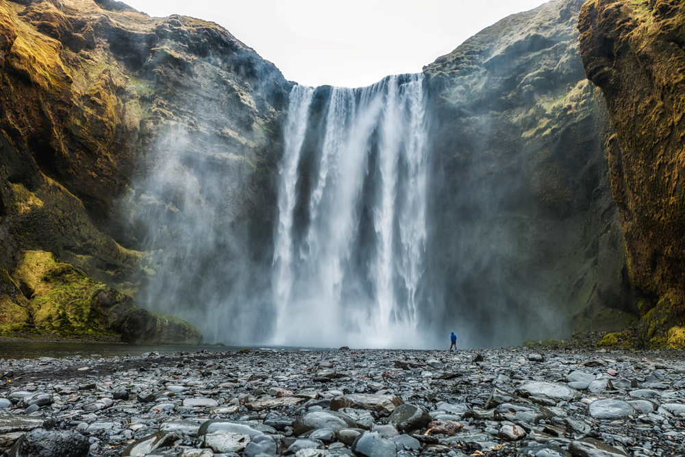 The largest waterfall in Iceland towers above the land while a tourist walks by it.