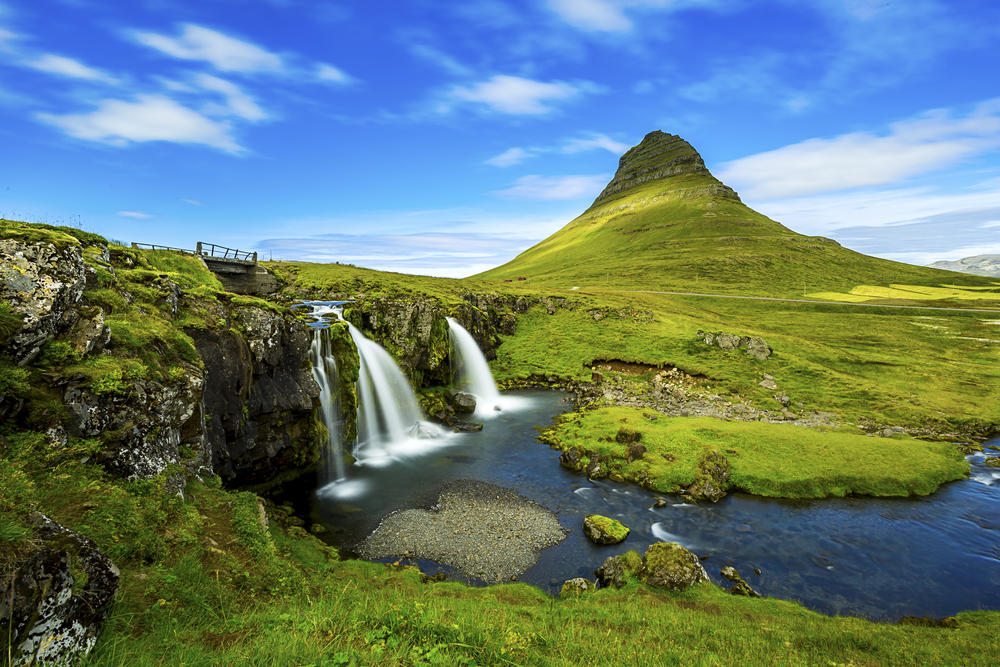 Kirkjufell Mountain towers high over the land in green with a blue sky behind it in west iceland
