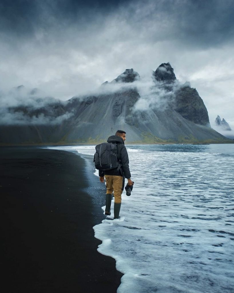 Man with backpack and camera walking at the edge of the water at Stokksnes bundled up for winter with misty mountains in the background.