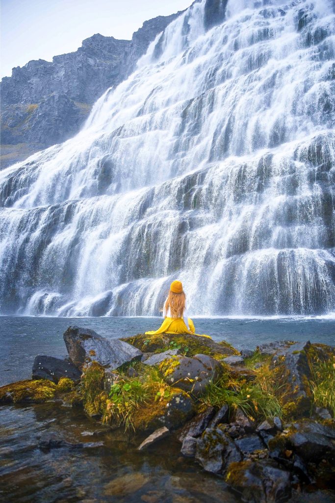 A woman in a yellow hat and skirt sitting in front of the Dynjandi waterfall in the Westfjords in Iceland in May