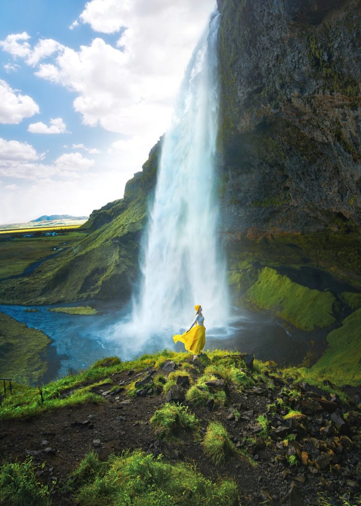 A woman standing on a rock wearing a yellow hat and yellow skirt blowing in the wind, beside the walking path around the Seljalandsfoss waterfall