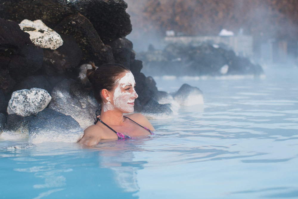 A woman relaxes in the blue lagoon with mud on her face.