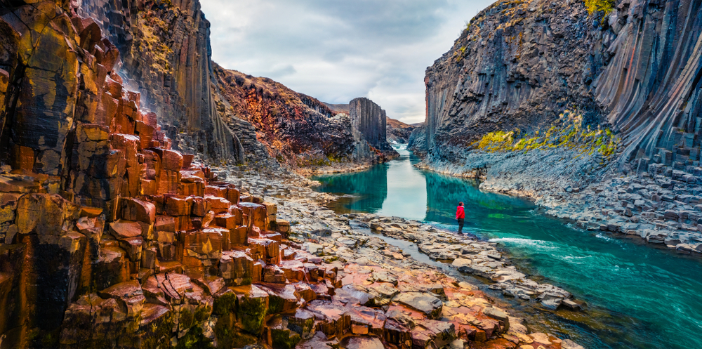 A tourist in a red jacket overlooks the blue waters in an Icelandic canyon.
