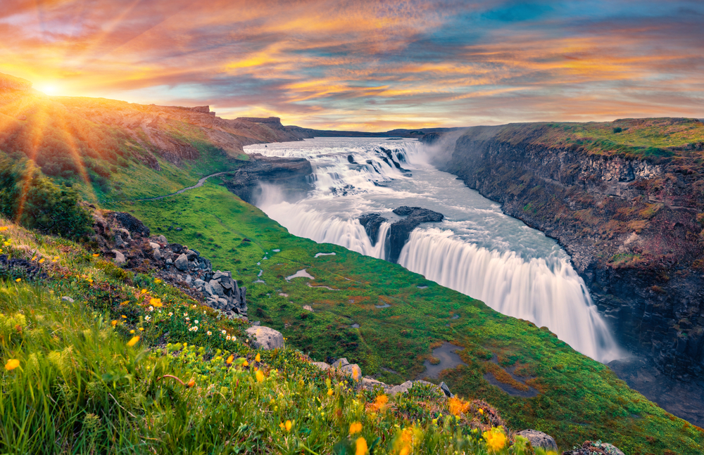 a summertime view of Gullfoss waterfall with green grass, wildflowers, and the sun shining overhead