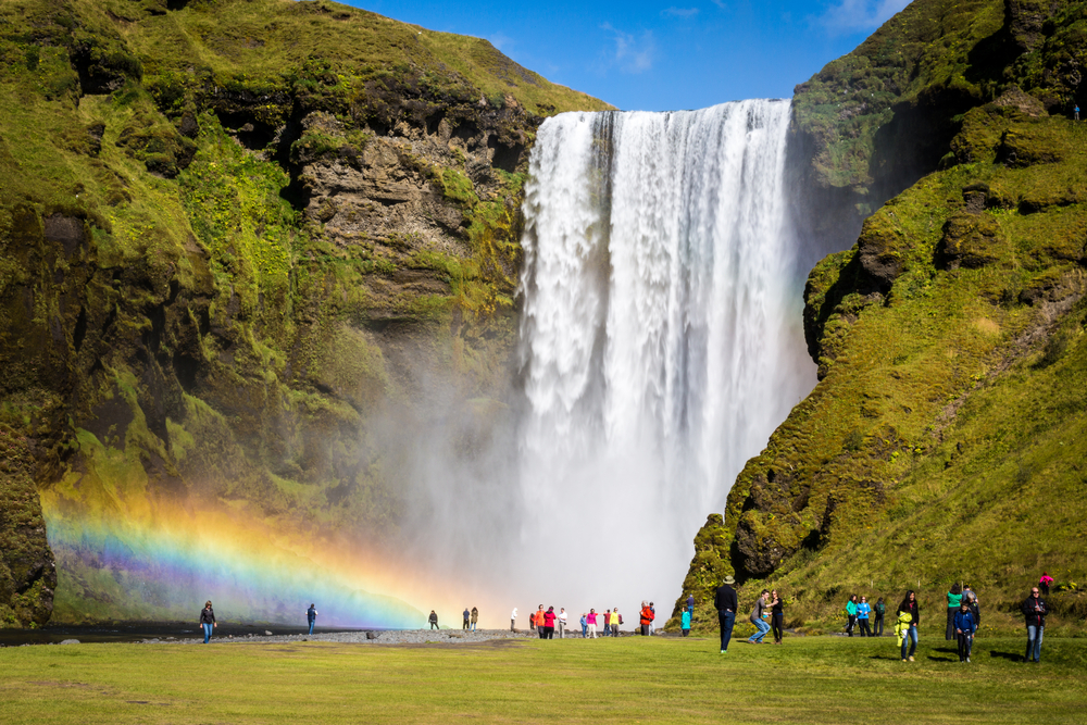 A sunny August day at Skogafoss Waterfall with a rainbow and groups of tourists.