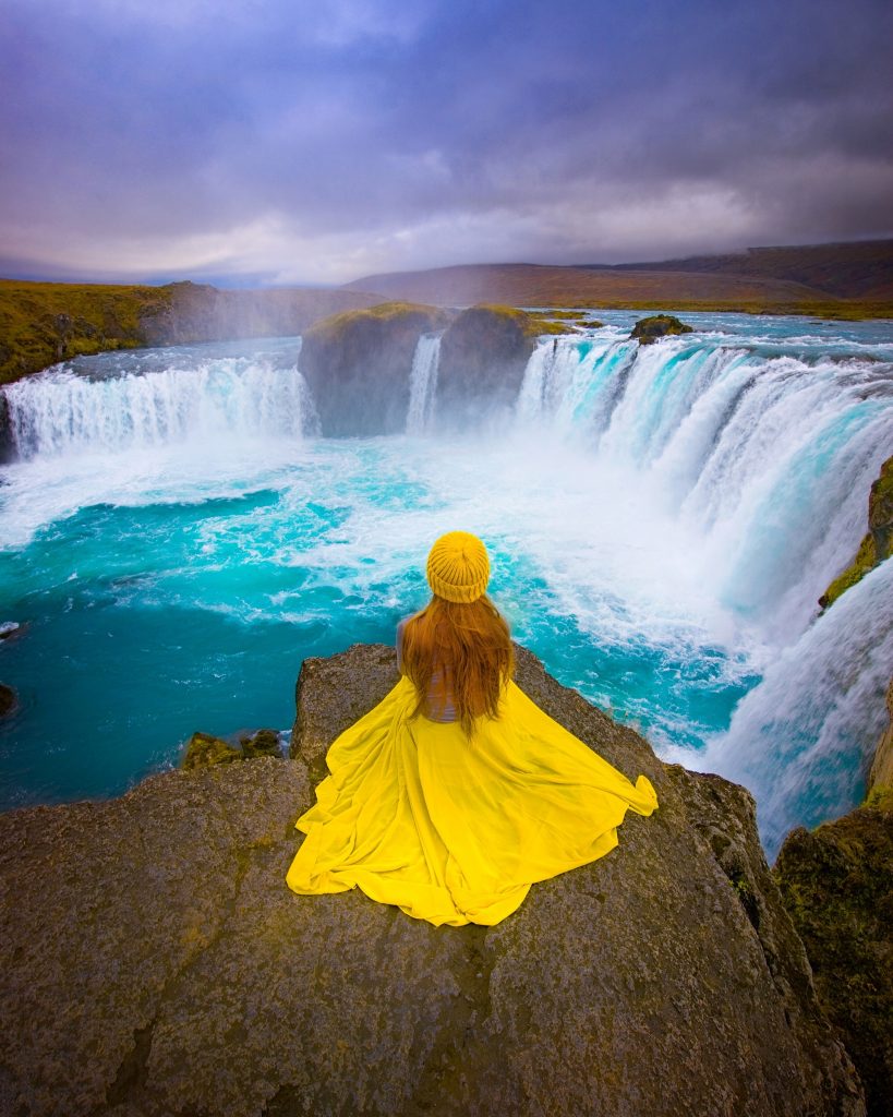 Girl in yellow dress and hat sits on a cliff edge overlooking the blue water of Godafoss Waterfall.