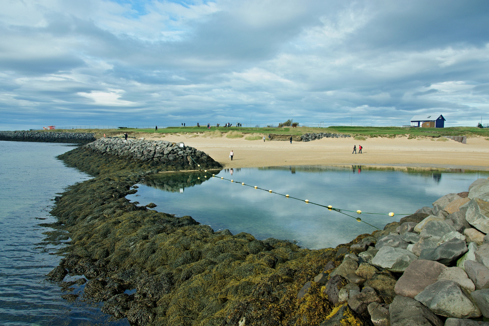 Looking across the water towards Nautholsvik Beach where people walk on the sand.