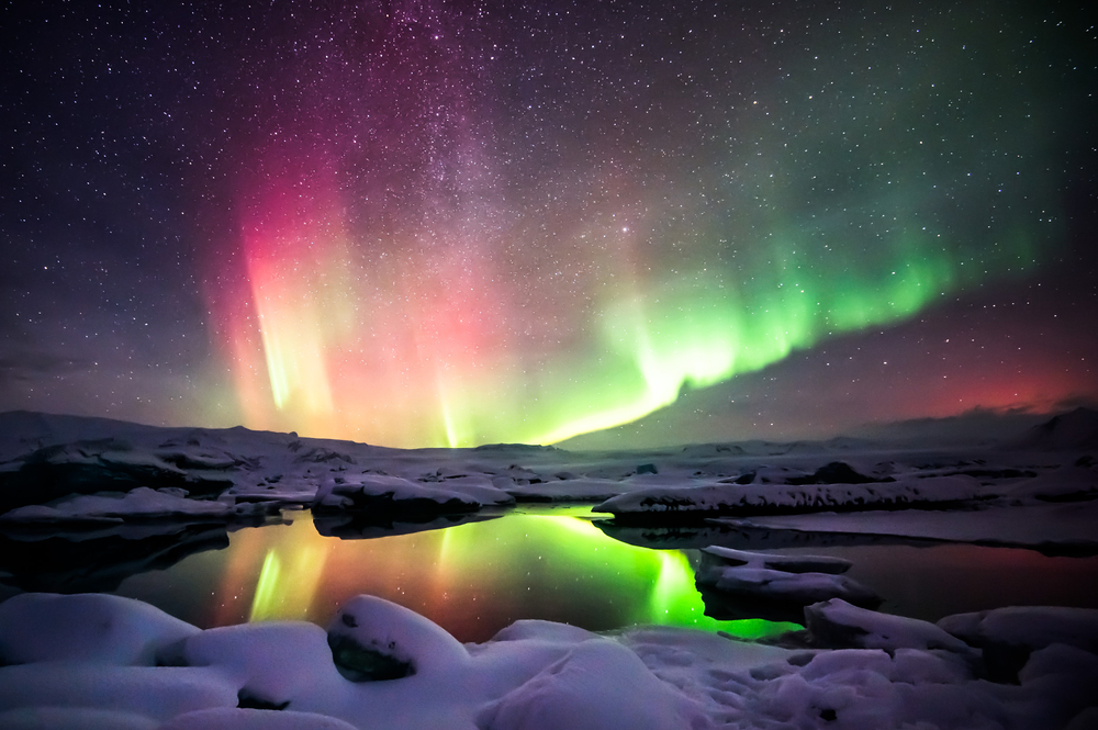 The dazzling pink and green colors of the Northern Lights and stars reflecting in a glacier lagoon.