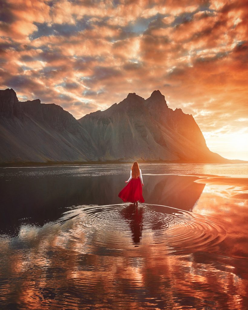Girl in red skirt walks through the tide toward Stokksness during a bright, orange sunrise.