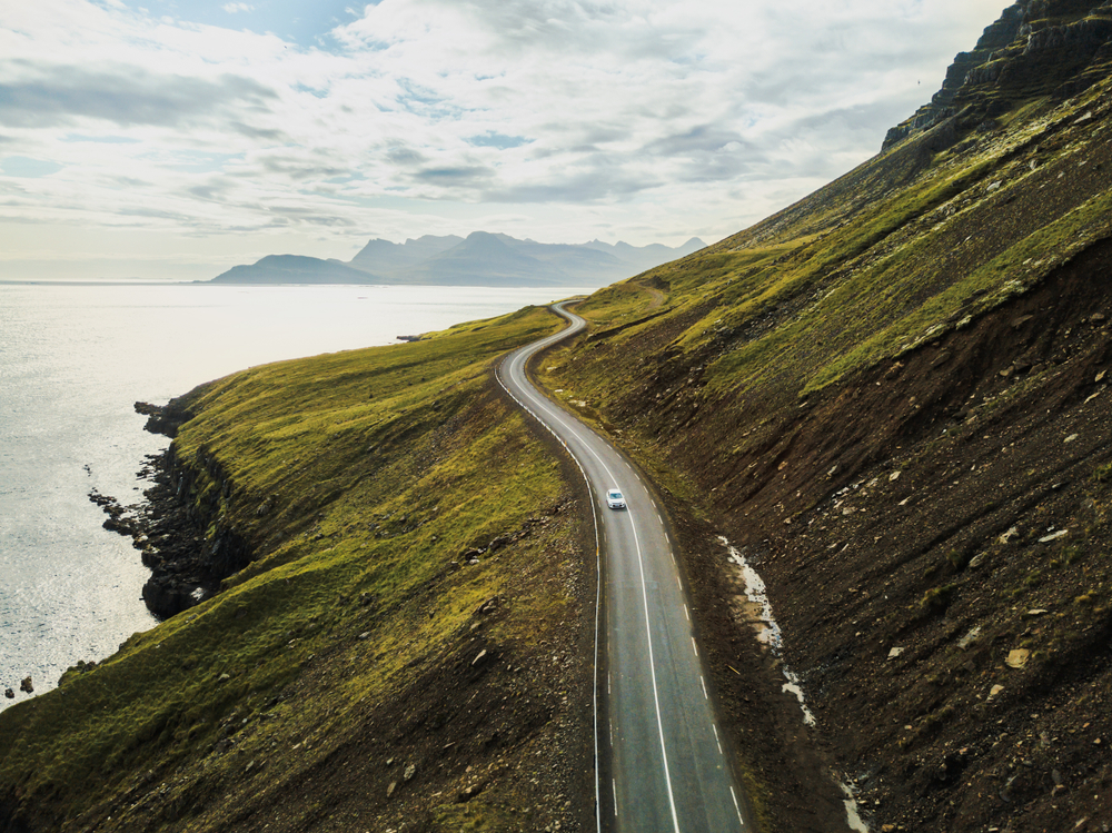 a single car driving a paved road with the ocean to one side and a mountain to the other