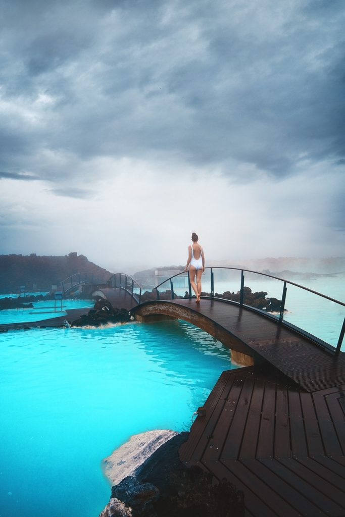 Woman in white swim suit walks across a bridge over the pretty blue, steaming water of the Blue Lagoon, one of the best hot springs in Iceland.