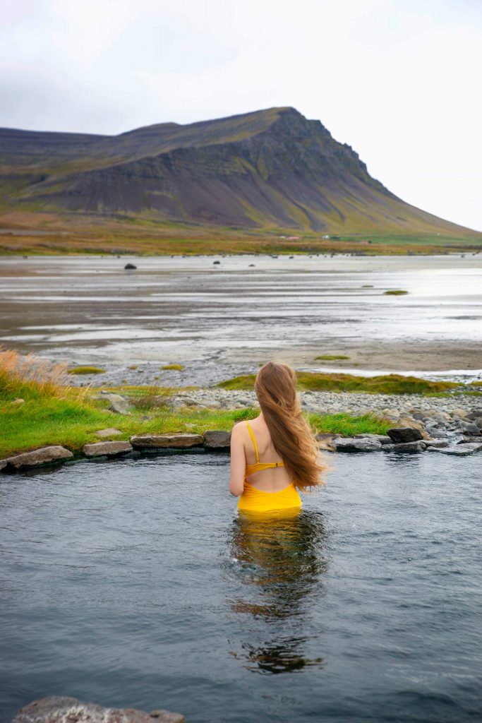 Woman in a yellow swim suit stands in the Krosslaug Hot Springs looking out at a distance mountain across the water.