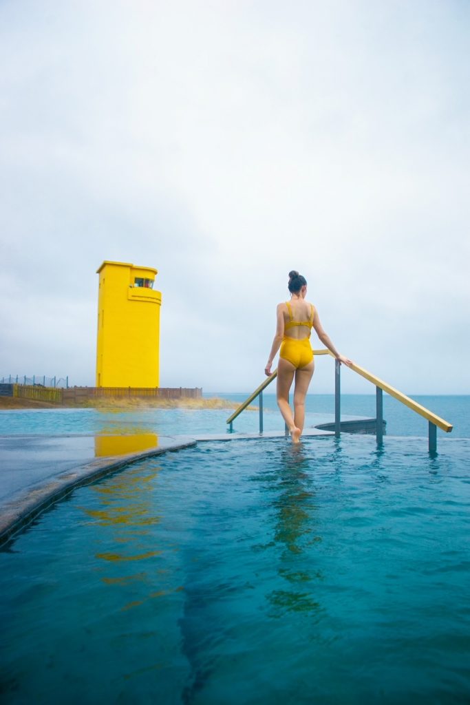 Woman in a yellow swimsuit walks up steps in a hot spring near a yellow lighthouse.