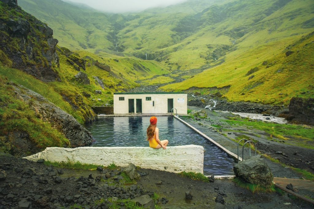 Woman in yellow swim suit and hat sits on the edge of the Seljavallalaug Hot Springs on an overcast day,