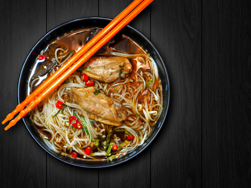 a black bowl of noodle soup with yellow chopsticks and chicken thighs 