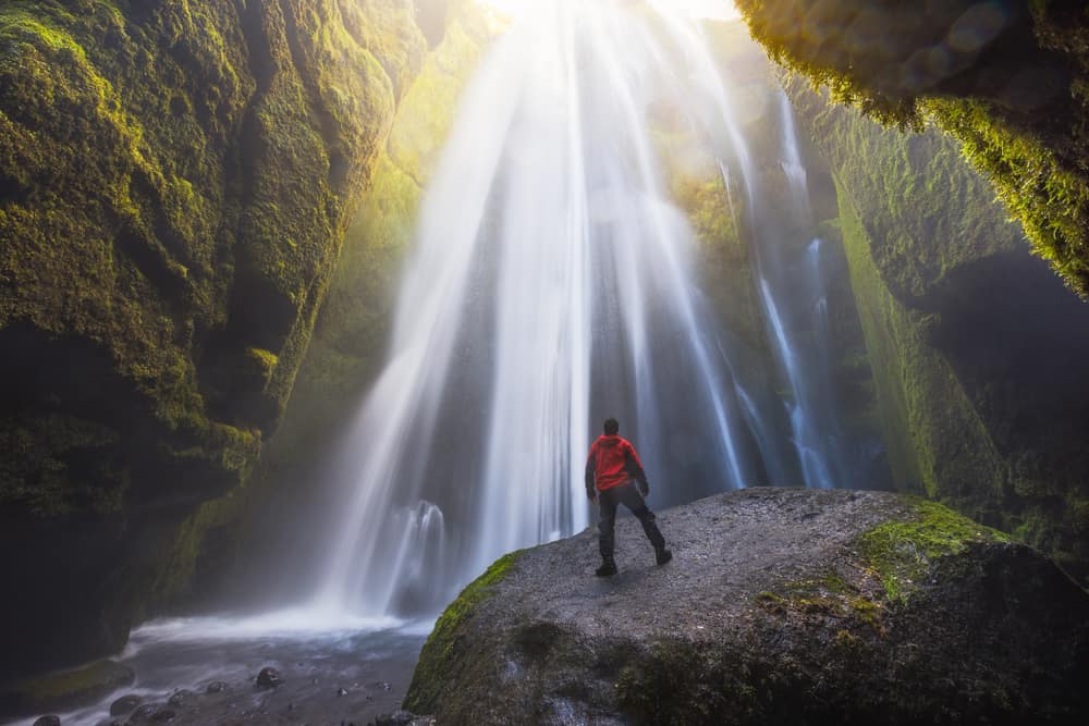 a guy standing under a waterfall with rocks on both side and sun shining fro top