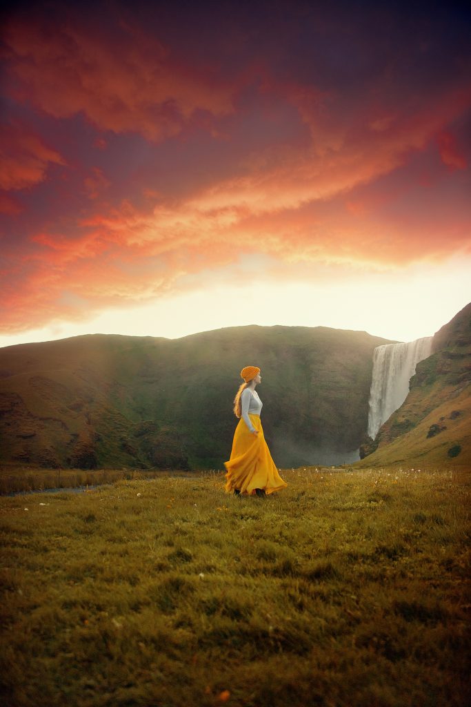 a girl in a yellow skirt in front of a waterfall at sunset in green grass