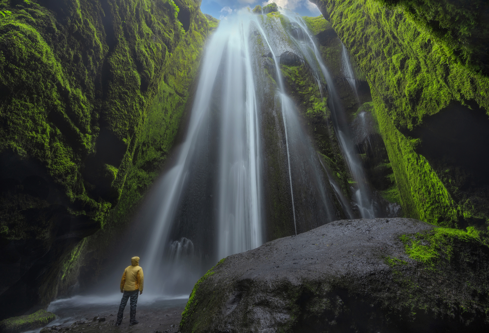 A person with a yellow rain jacket standing in the basin of a waterfall cascading down a mossy cliff side