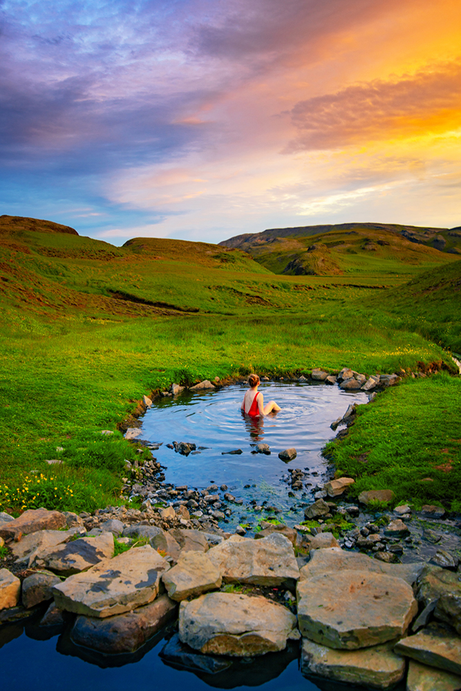 A colorful sunset over Hrunalaug Hot Spring with a girl in a red swimsuit sitting in a round pool among green hills.
