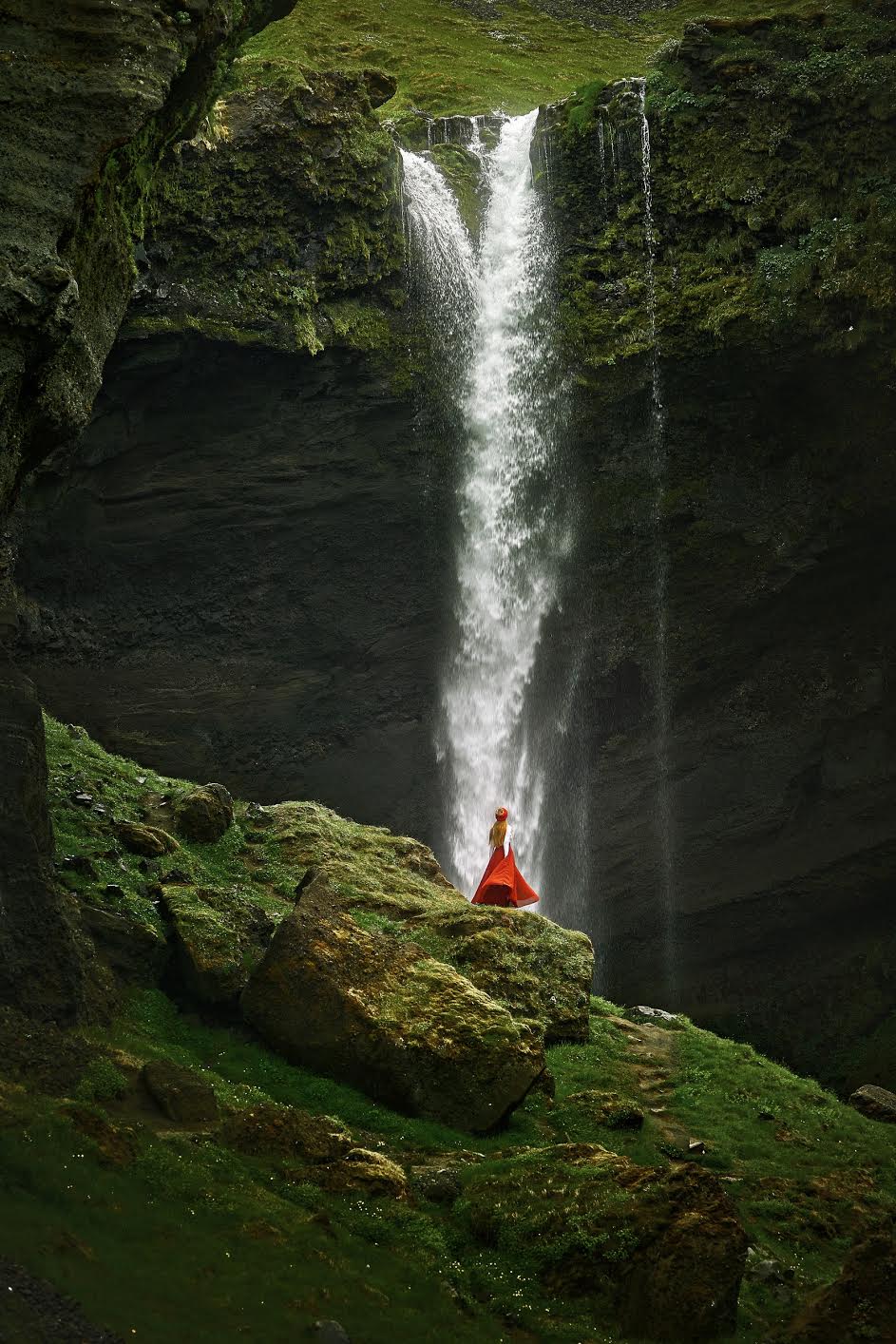 A person in a red skirt standing on a mossy rock looking out at a massive waterfall that is falling off the side of a cliff. 