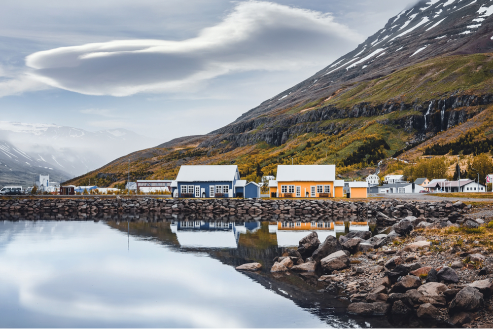 A cloud looms over a blue and yellow colorful houses beside reflective lake with moss covered mountain in the background once where famous movies filmed in Iceland.