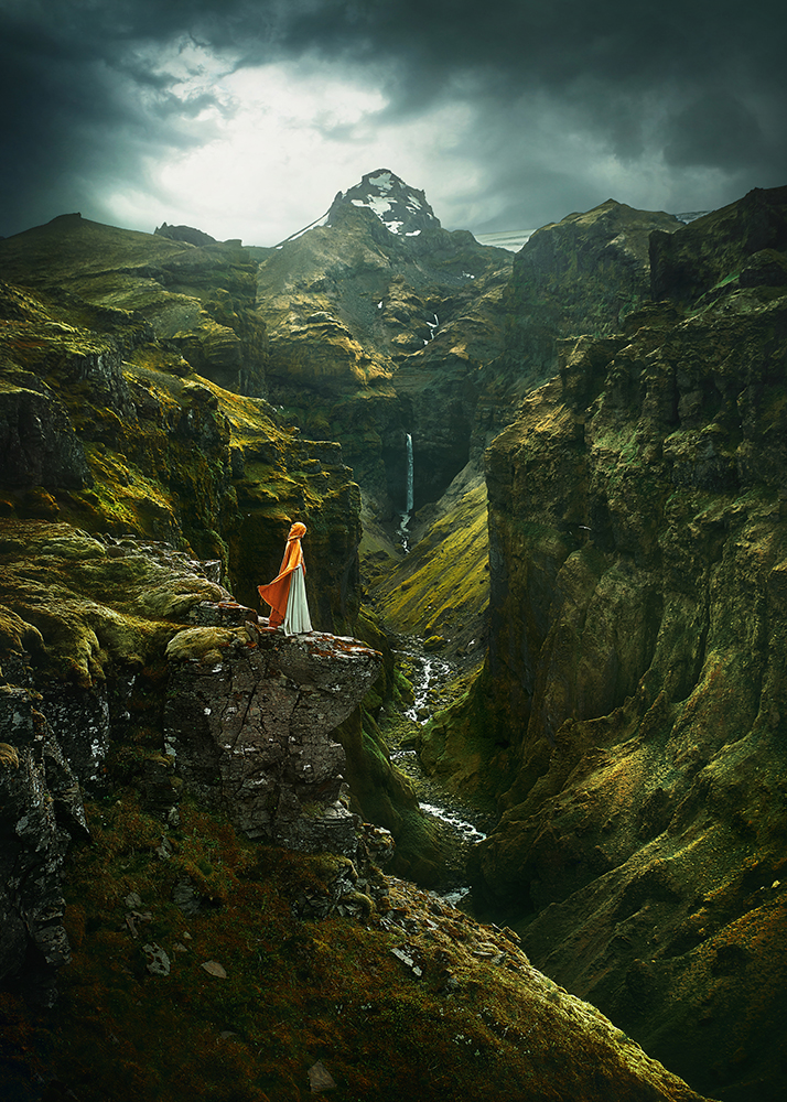 Woman in yellow cape stands on the edge of Mulagljufur Canyon with rugged mountains and a waterfall in the distance.