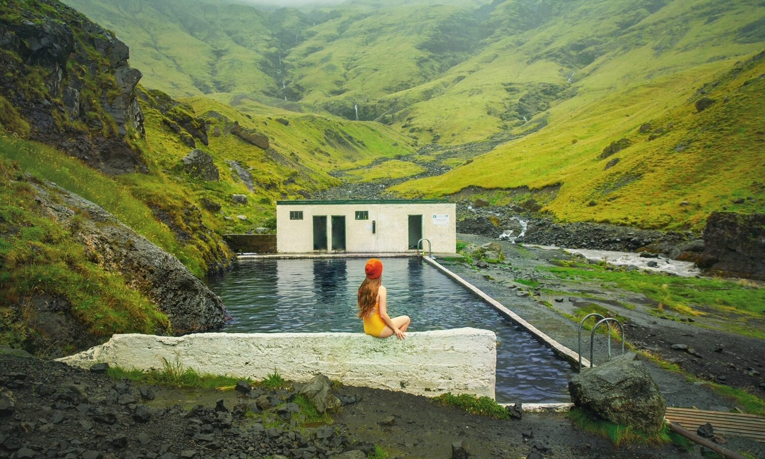 woman sitting in yellow swimsuit on the edge of a hot spring in iceland with green mountains in the background