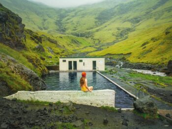 woman sitting in yellow swimsuit on the edge of a hot spring in iceland with green mountains in the background